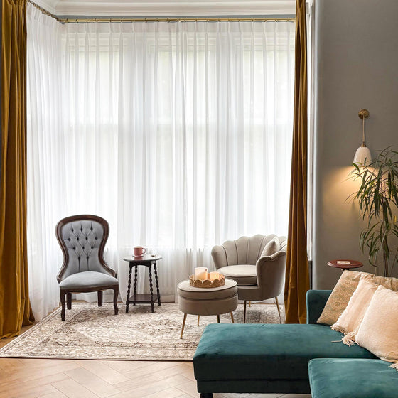 Weiß, Wohnzimmer mit blaugrünem Sofa, grauem Sessel und beigem Sessel neben großen Fenstern mit weißen transparenten und goldenen Vorhängen.