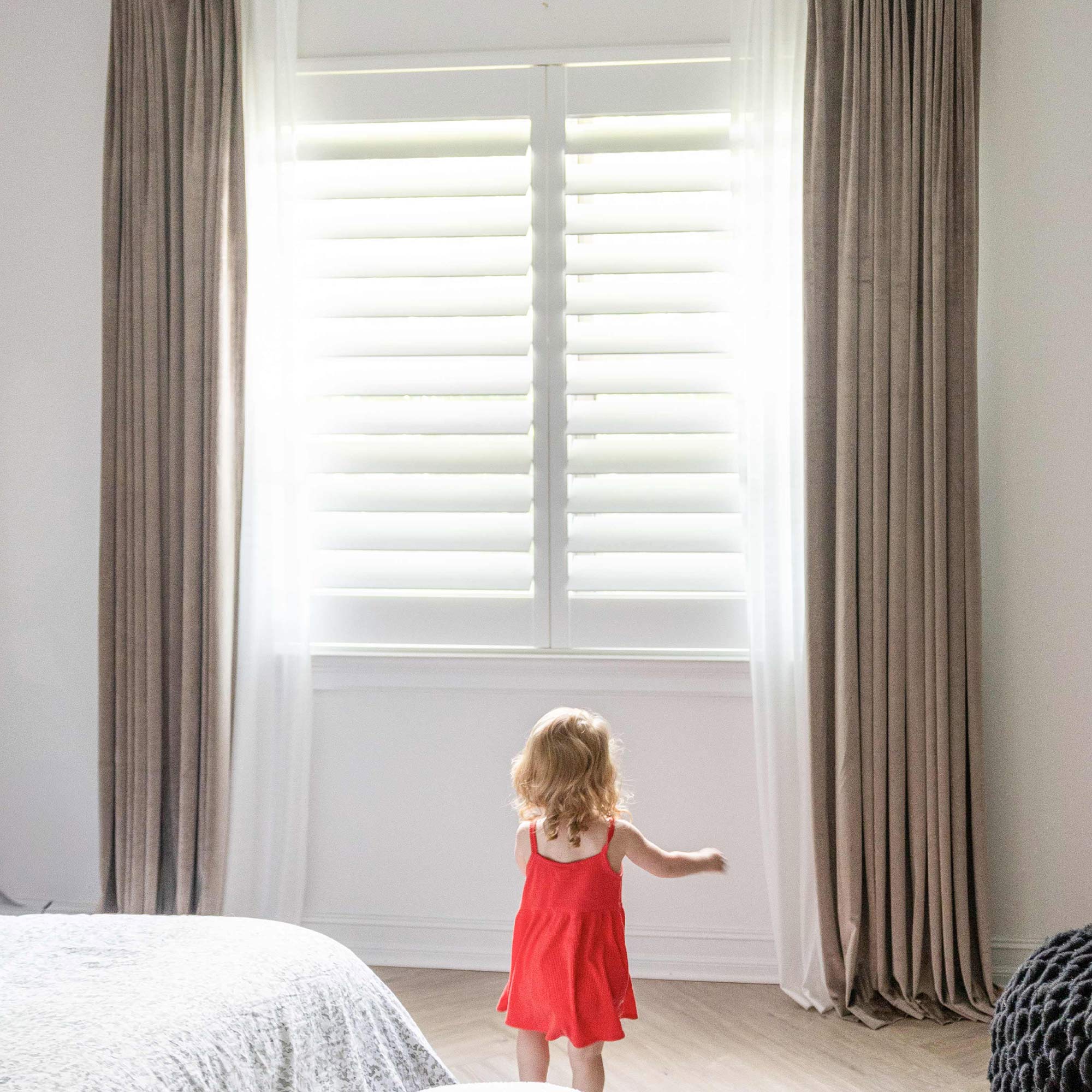 Taupe, Kind in rotem Kleid vor einem Fenster mit weißen Fensterläden und beigen Vorhängen.