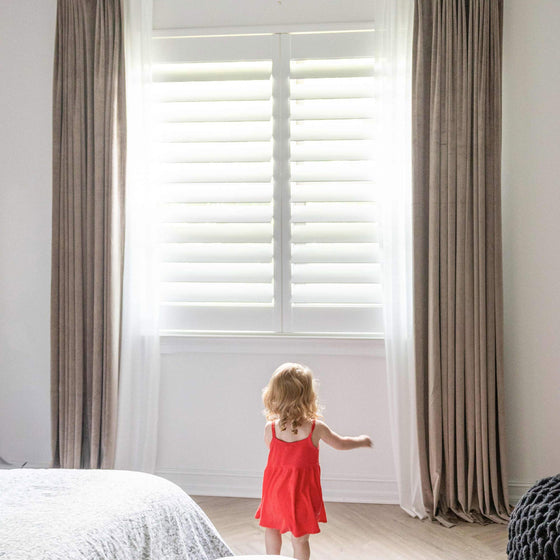 Taupe, Kind in rotem Kleid vor einem Fenster mit weißen Fensterläden und beigen Vorhängen.